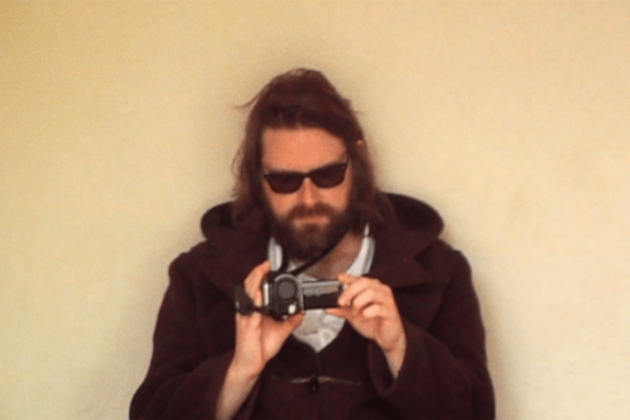 Bitw (Gruff ab Arwel) in sunglasses and a duffel coat, filming with a camcorder against a cream wall