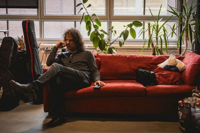 Martin Saint, with long curly hair and dressed in black, sitting on a red sofa in a studio with guitar cases and pot plants