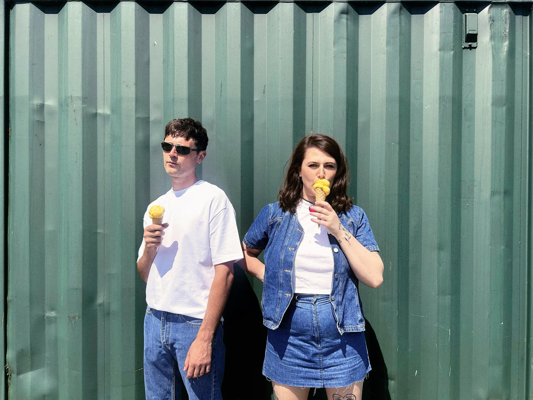 British Birds - Bobby and Emma Townson in white T-shirts and denim, eating ice creams in front of a green corrugated metal wall