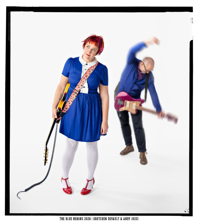 The Blue Herons: Gretchen DeVault in a blue dress and red shoes holding a sunburst guitar, with Andy Jossi blurred behind her swinging a pink Jazzmaster-style guitar
